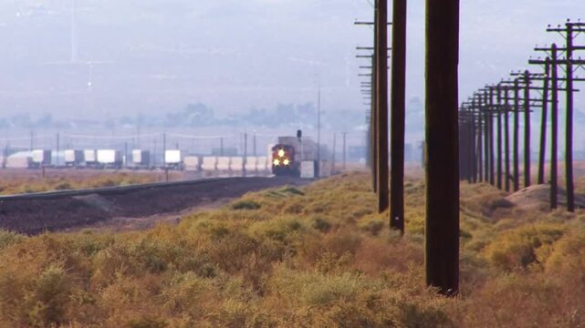Long shot of an approaching double-stack container freight train crossing a windy, weedy  field along a double line of telephone poles.