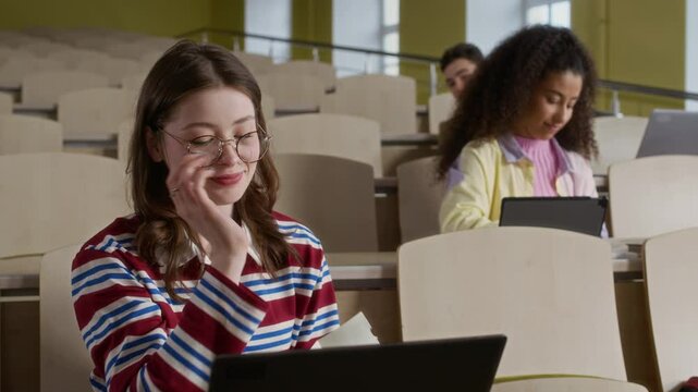 Medium shot of diverse peers sitting at tiered desks giving secret note to gen Z friend having class in college