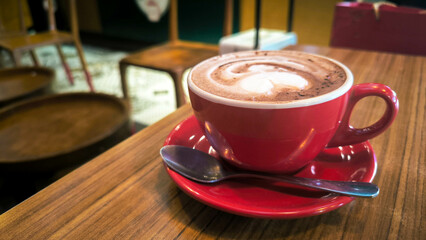 A cup of hot chocolate or coffee with latte art served in a red cup on a wooden café table, accompanied by a spoon.