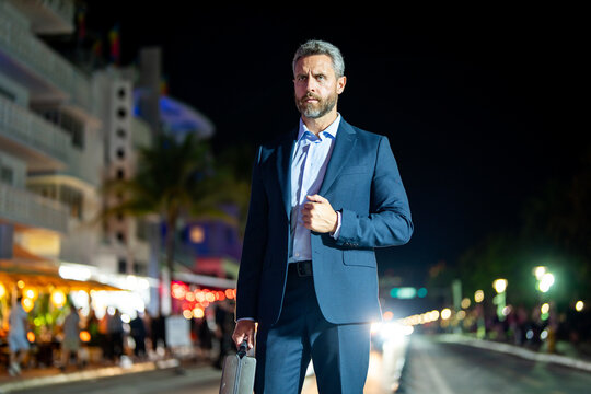 Businessman with briefcase walking at night city. Rich man in suit in city. Businessman near luxury car. Businessman walking near night city lights. Business style. Rich businessman in luxury setting.