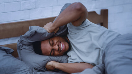 African American Man Suffering From Headache And Insomnia, Covering Ears With Pillow Hearing Voices...