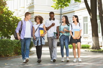 Fototapeta premium Group of multiethnic students walking together outdoors in college campus, holding books and notepads and laughing, copy space