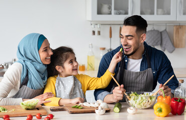 Taste It. Portrait of cute little arab girl feeding her daddy with cucumber slice, happy middle eastern family of three cooking healthy tasty food in kitchen together, making vegetable meal at home
