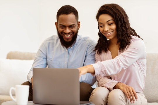 Cheerful african american couple relaxing together at home, using laptop, choosing video or movie. Happy black man and woman sitting with computer on the couch in cozy living room or bedroom - Powered by Adobe