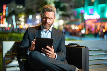 Online business. Mature business man working on tablet outdoors. Business man with tablet outdoor. Businessman with tablet on night street. Businessman using tablet, sit on bench. Night business.