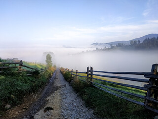 Rural mountain path lined with wooden fences disappearing into thick morning fog.