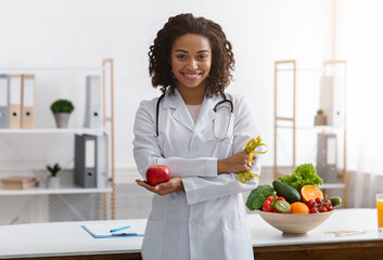 African female dietician with crossed arms holding fresh apple, posing at workplace