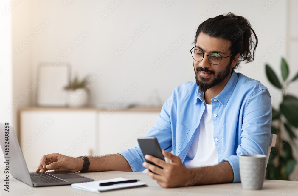 Poster millennial arab freelancer guy using smartphone while working on laptop at desk in home office, brow