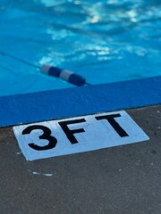 a painted pool edge with a 3 foot depth sign  on the sidewalk - corner of swimming pool, blue water and a safety rope. High angle view of public swim park