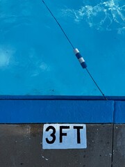 The shallow end of a pool with a 3 foot depth sign painted on the concrete - edge of swimming pool, blue water and a safety rope. High angle view of public swim park