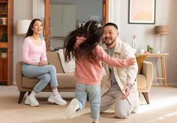 Reunion With Daddy. Asian Kid Daughter Running To Embrace Her Dad, While Mother Sitting On Sofa In Modern Living Room At Home. Family Celebrating Father's Homecoming. Selective Focus