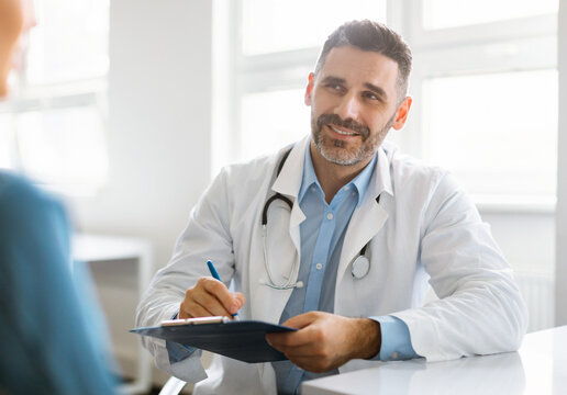 Cheerful middle aged male doctor in white coat with stethoscope consulting patient in office clinic interior. Medical consultation, diagnostics, therapy, business and health care