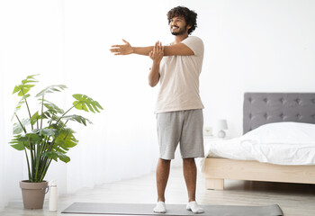 Cheerful millennial indian man in homewear practicing yoga at home, standing on fitness mat and...