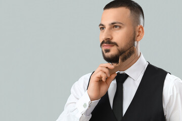 Handsome young bearded man combing beard on grey background