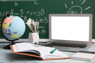 Notebook with calculator, laptop and globe on table near blackboard, closeup. Student loan concept
