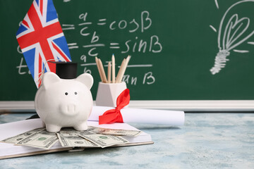 Piggy bank with money, diploma, book and UK flag on table near blackboard. Student loan concept