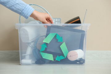 Female hand with electronic waste for recycling in container near beige wall