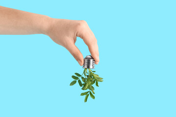 Fototapeta premium Female hand holding light bulb with plant leaves on blue background. Green technology concept