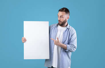 Surprised millennial man pointing at white blank poster on blue studio background, mockup for your...