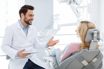 Male dentist showing female patient jaw x-ray on digital tablet, dental clinic interior. Bearded man stomatologist having conversation with lady in dental chair, holding pad, side view, copy space