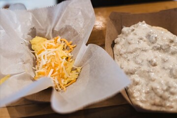 Takeout tray with fluffy scrambled eggs topped with shredded cheese alongside buttermilk biscuits smothered in creamy sausage gravy, close-up breakfast photo