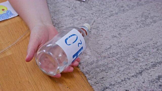 A person's hand rests on a light wooden floor gripping a clear plastic bottle labeled HO2 with a connected breathing tube next to a soft grey rug in a home interior dachshund dog runs up and sniffs