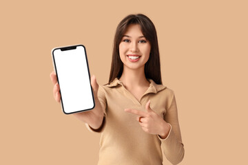 Smiling young woman pointing at blank mobile phone on beige background