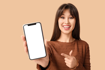 Smiling young woman pointing at blank mobile phone on beige background