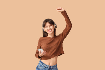 Smiling young woman in headphones with mobile phone dancing on beige background