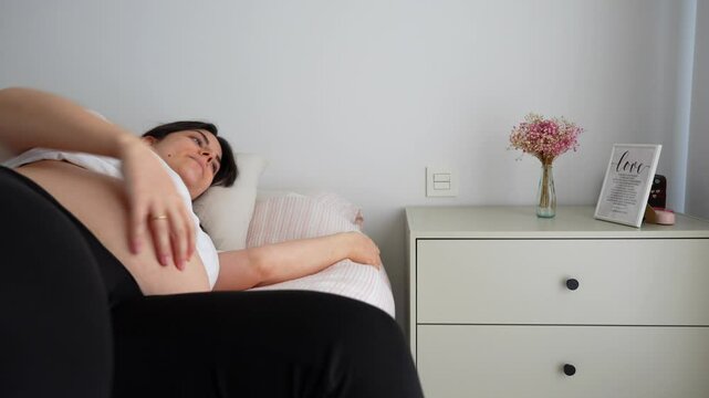 Pregnant woman in bed struggling to get up. Soft light, lactation pillow nearby