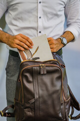 A man in a white shirt is putting a large book into a brown leather backpack.