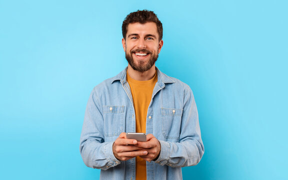 Cheerful guy with smartphone looking at camera and smiling, texting in social media over blue studio background. Positive young man in casual chatting on phone, using new mobile application