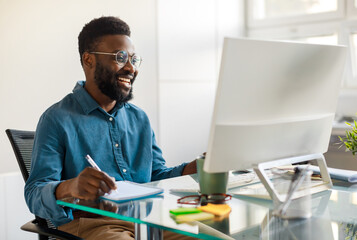 Positive black businessman writing notes in notebook while watching webinar video course, looking...