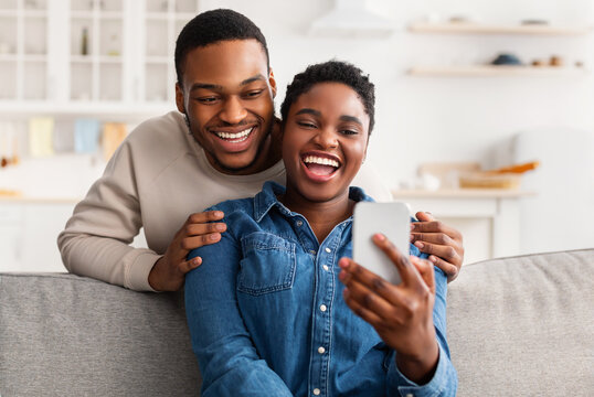 Smiling young African American man embracing his woman from behind while looking at smartphone. Loving couple sharing social media on mobile phone, happy female sitting on sofa, showing gadget - Powered by Adobe