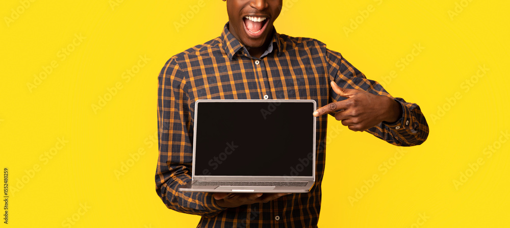 Poster Check This Website. Joyful African Guy Pointing At Laptop Computer With Black Blank Screen, Standing Isolated Over Yellow Background, Demonstrating Free Place For Your Design, Mockup With Copy Space