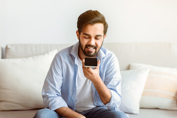 Smiling Arab Man Recording Voice Message On Smartphone Or Using Virtual Assistant While Relaxing On...