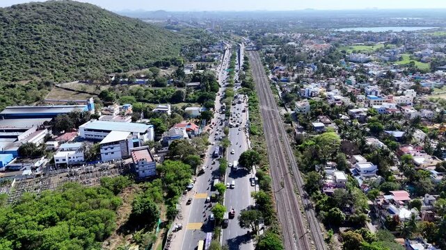 Aerial view of a major Chennai&ndash;Trichy highway (GST Road) and railway tracks in Chennai, flanked by a green hill on one side and urban development on the other, under a clear sky.