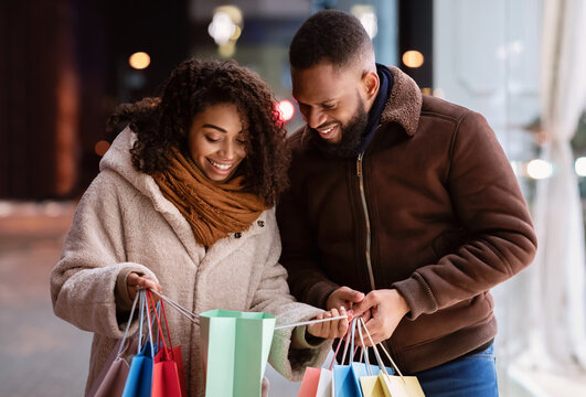 Portrait of lovely black couple holding many colorful shopping bags, looking inside, woman showing what she bought to her curious boyfriend or husband, family walking down the street in the evening