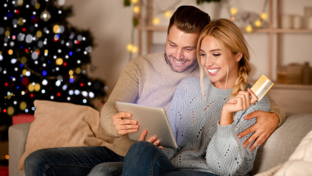 Delighted husband and wife making purchases online, using digital tablet and credit card, Christmas atmosphere at home. Happy young family shopping online on pad, using Christmas offers, copy space