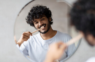 Toothcare routine concept. Young indian guy cleaning teeth in the morning using toothbrush and...