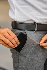 A man's hand with a black leather belt is putting a black leather wallet into his trouser pocket.