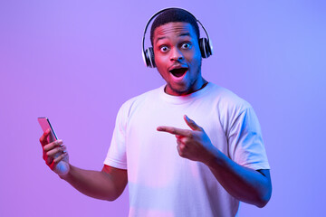 Mobile Offer. Surprised African Guy In Headphones Pointing At Smartphone In His Hand, Excited Black Man Recommending New App, Standing In Vivid Neon Light Over Purple Background, Looking At Camera