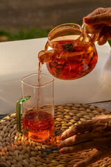 Close-up of woman's hand pouring strawberry tea from glass teapot into glass mug. Vertical...