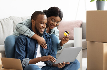 Online Shopping Concept. Cheerful Black Spouses With Laptop Choosing Furniture After Moving To New...