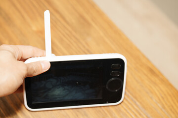 A parent’s hand placing a baby monitor on a wooden table. The baby monitor has a white frame, visible antenna, and dark screen displaying a dimly lit image from the baby’s room