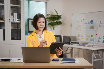 A middle-aged Asian woman sits at a work desk, deeply focused. Surrounded by charts and notes, she analyzes data, plans strategies, and prepares to lead a presentation or consultative session