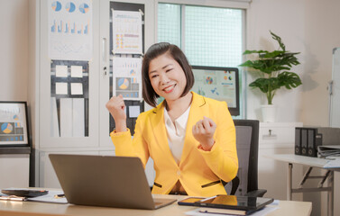 A middle-aged Asian woman sits at a work desk, deeply focused. Surrounded by charts and notes, she...