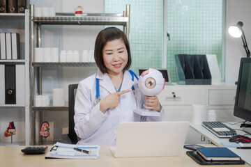 A middle-aged Asian female doctor holds an eye model at her desk, explaining eye anatomy and vision problems.She educates patients or, demonstrating knowledge as an optometrist or medical instructor