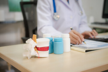 A middle-aged Asian female doctor sits at her desk, pointing to a spinal model with a highlighted...