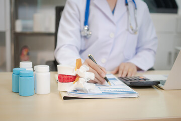A middle-aged Asian female doctor sits at her desk, pointing to a spinal model with a highlighted...